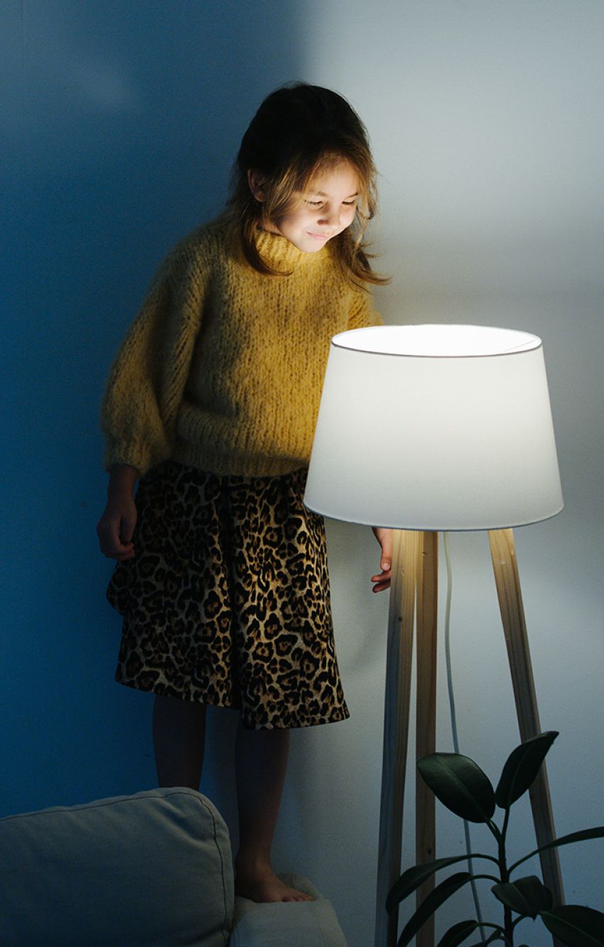 Brunette little girl in yellow knitted sweater and leopard skirt standing on a backseat of a couch at home. She's looking at dim night light from above. Her face is lit. 