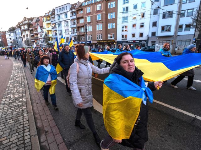 Menschen ziehen in einer Demonstration durch eine Stadtstraße und tragen eine große blau-gelbe Flagge.