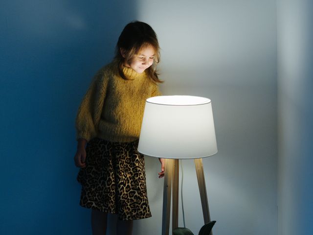 Brunette little girl in yellow knitted sweater and leopard skirt standing on a backseat of a couch at home. She's looking at dim night light from above. Her face is lit. 