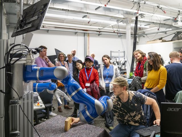 Eine Gruppe von Teilnehmenden mit blauen Namensbändern beobachtet in einem Robotiklabor der TU Darmstadt eine Demonstration, bei der ein blau-weißer kollaborativer Roboterarm einen Holzklotz greift, während ein junger Mann mit Brille und Laptop die Steuerung übernimmt. 