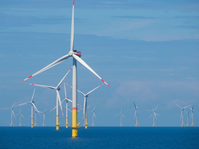 Mehrere Offshore-Windräder stehen im Meer vor blauem Himmel, mit einer großen Turbine im Vordergrund. 