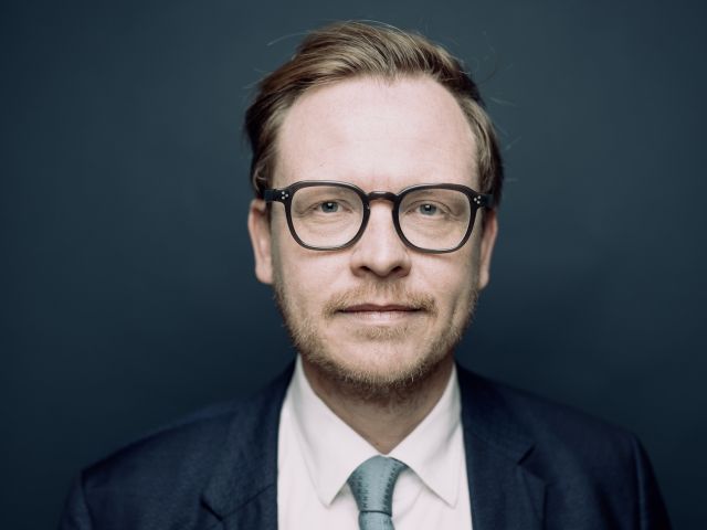 Porträt eines Mannes mit runder schwarzer Brille, kurzem Bart und Seitenscheitel, der in Anzug, weißem Hemd und hellgrüner Krawatte vor dunkelblauem Hintergrund in die Kamera blickt. 