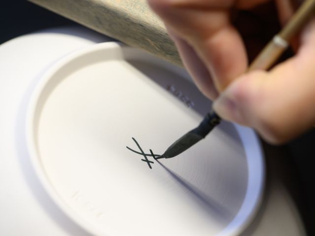Eine Hand hält einen Pinsel und malt auf die Unterseite einer Schale zwei gekreuzte Schwerter.
