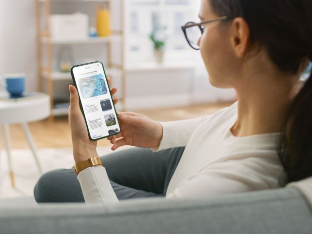 Young Woman at Home Uses Smartphone for Scrolling and Reading News about Technological Breakthroughs. She's Sitting On a Couch in Her Cozy Living Room. Over the Shoulder Shot