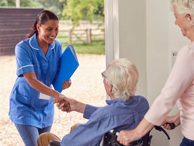 Senior Couple With Man In Wheelchair Greeting Female Nurse Or Care Worker Making Home Visit At Door