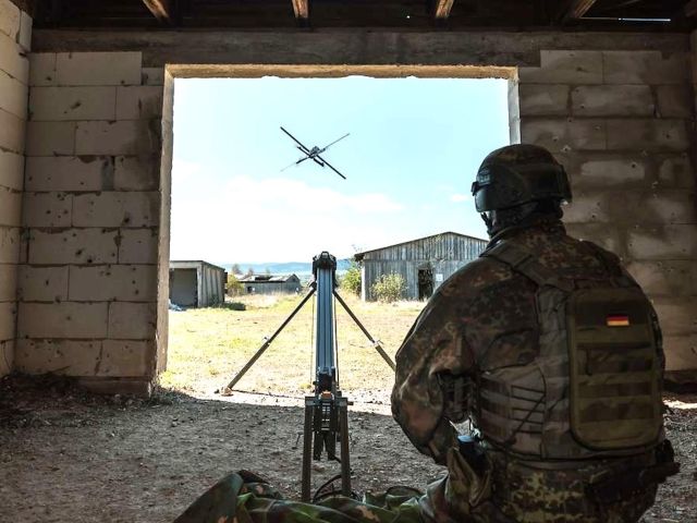 Soldat in Tarnuniform sitzt in einem Gebäude und steuert eine Drohne, die durch eine Maueröffnung ins Freie fliegt.