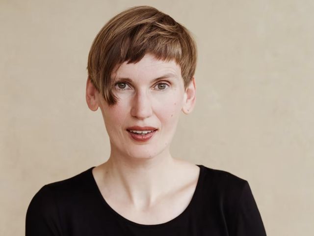 Porträt einer kurzhaarigen Person in schwarzem Oberteil vor hellem, unscharfem Hintergrund, die direkt in die Kamera blickt. 