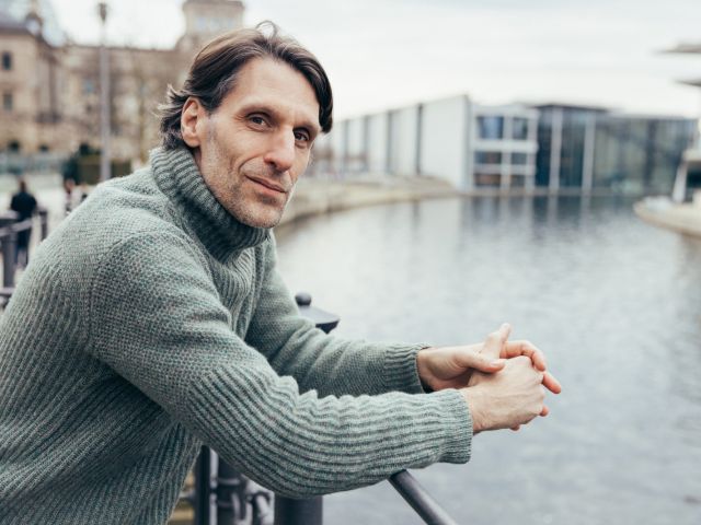 Mann in grauem Rollkragenpullover lehnt an einem Geländer am Flussufer und blickt in die Kamera, im Hintergrund sind Gebäude und ein modernes Bauwerk zu sehen. 