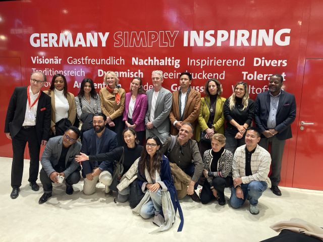 Gruppenfoto von etwa 16 Personen, die in zwei Reihen vor einer roten Wand mit dem Schriftzug „Germany Simply Inspiring" und weiteren Begriffen wie „Visionär", „Gastfreundlich" und „Nachhaltig" posieren. 