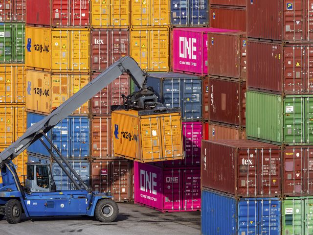 Ein blauer Containerstapler hebt einen gelben Frachtcontainer vor hohen Stapeln aus bunten Schiffscontainern im Hafen.