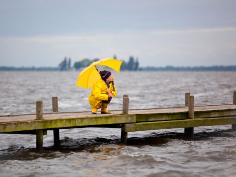 Junge Frau mit gelber Regenjacke und Schirm in der Hocke auf einem Steg 