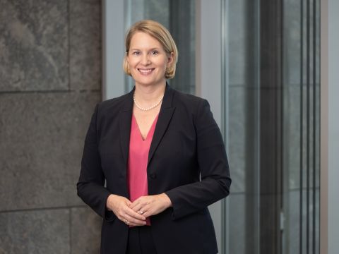 Professionelles Porträtfoto einer blonden Frau mit kinnlangem Haar, die einen schwarzen Blazer über einem roten Oberteil trägt und mit verschränkten Armen vor einem unscharfen grünen Hintergrund lächelt. 