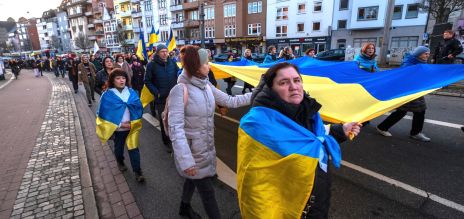 Personas participan en una manifestación por una calle de la ciudad mientras portan una gran bandera azul y amarilla.