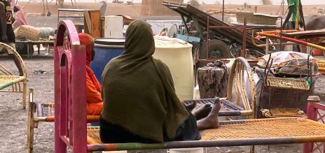 Una persona sentada sobre la estructura de una cama trenzada al aire libre. Detrás hay muebles dispersos, contenedores y un vehículo en un paisaje con mucho polvo.