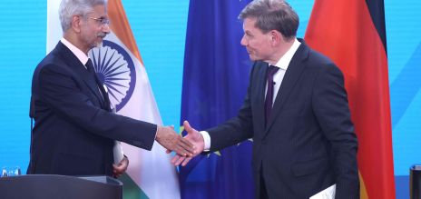 Two men in suits shake hands on a stage, with the national flags of India and Germany visible in the background, along with the flag of the European Union. 