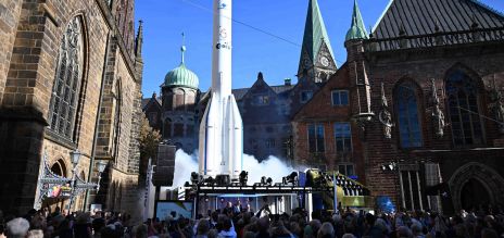 A white replica of an Arianne rocket on a stage in front of historic buildings 