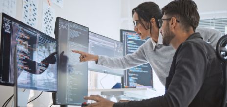 Man and woman in an office in front of multiple computer screens. 