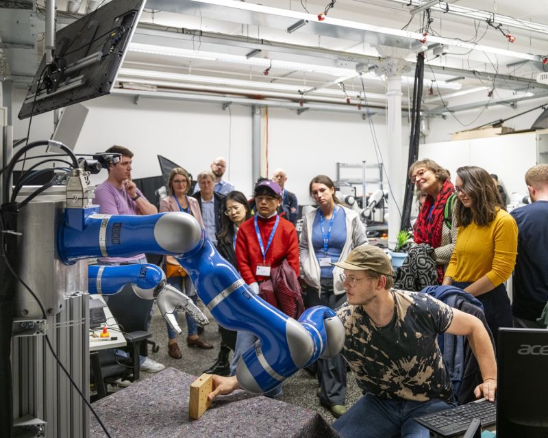 Eine Gruppe von Teilnehmenden mit blauen Namensbändern beobachtet in einem Robotiklabor der TU Darmstadt eine Demonstration, bei der ein blau-weißer kollaborativer Roboterarm einen Holzklotz greift, während ein junger Mann mit Brille und Laptop die Steuerung übernimmt. 