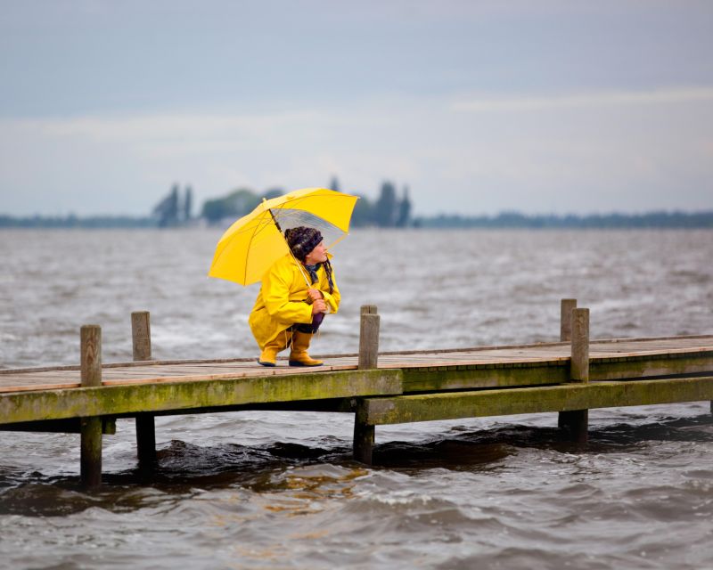 Junge Frau mit gelber Regenjacke und Schirm in der Hocke auf einem Steg 