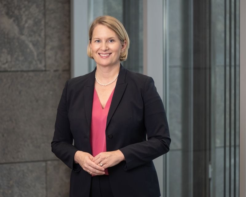 Professionelles Porträtfoto einer blonden Frau mit kinnlangem Haar, die einen schwarzen Blazer über einem roten Oberteil trägt und mit verschränkten Armen vor einem unscharfen grünen Hintergrund lächelt. 