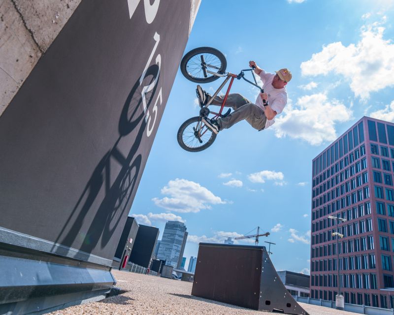 BMX-Fahrer springt mit dem Rad an einer Wandrampe hoch; im Hintergrund moderner Bürobau unter blauem Himmel.