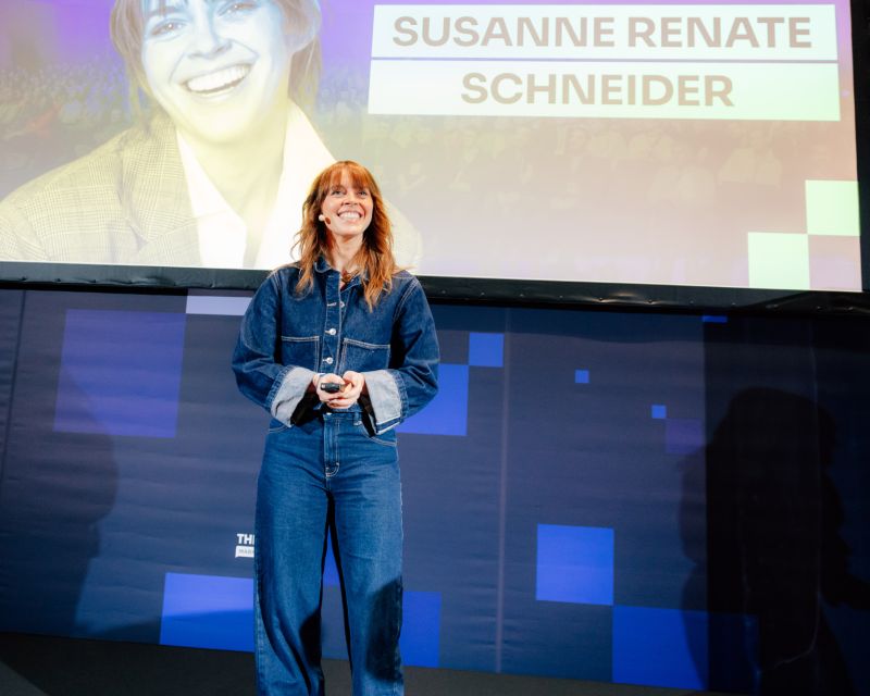 Eine lächelnde Frau mit Headset-Mikrofon steht auf einer Bühne und hält eine Fernbedienung, während hinter ihr auf einer großen Leinwand der Name „SUSANNE RENATE SCHNEIDER“ angezeigt wird.