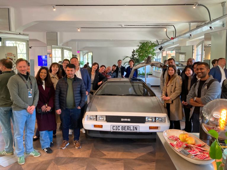 Eine Gruppe von Menschen posiert in einem hellen Innenraum um die Front eines DeLorean-Autos, dessen Kennzeichen „CIC BERLIN“ zeigt.