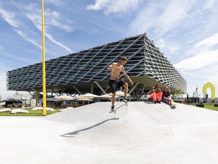 Ein Mann springt mit einem Skateboard über eine Betonrampe mit futuristischem Gebäude im Hintergrund