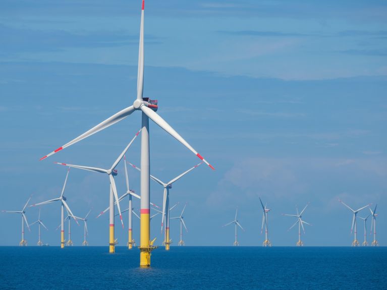 Mehrere Offshore-Windräder stehen im Meer vor blauem Himmel, mit einer großen Turbine im Vordergrund. 