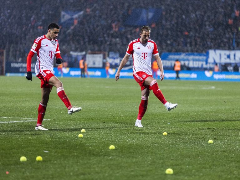 Football players Jamal Musiala and Harry Kane on a football pitch peppered with tennis balls. 