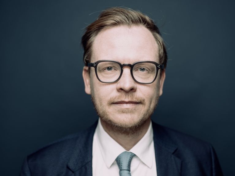 Portrait photo of a man with round black glasses, a short beard and side parting, wearing a suit, white shirt and light-green tie against a dark-blue background and looking into the camera. 