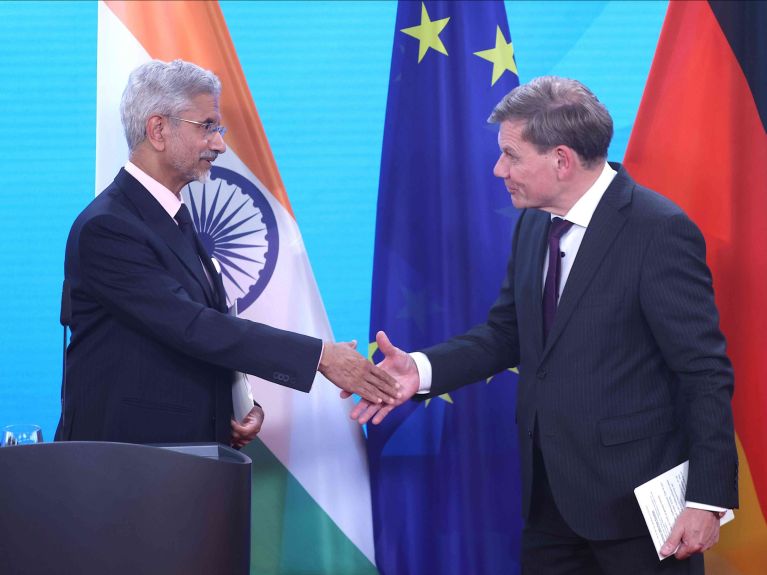 Two men in suits shake hands on a stage, with the national flags of India and Germany visible in the background, along with the flag of the European Union. 