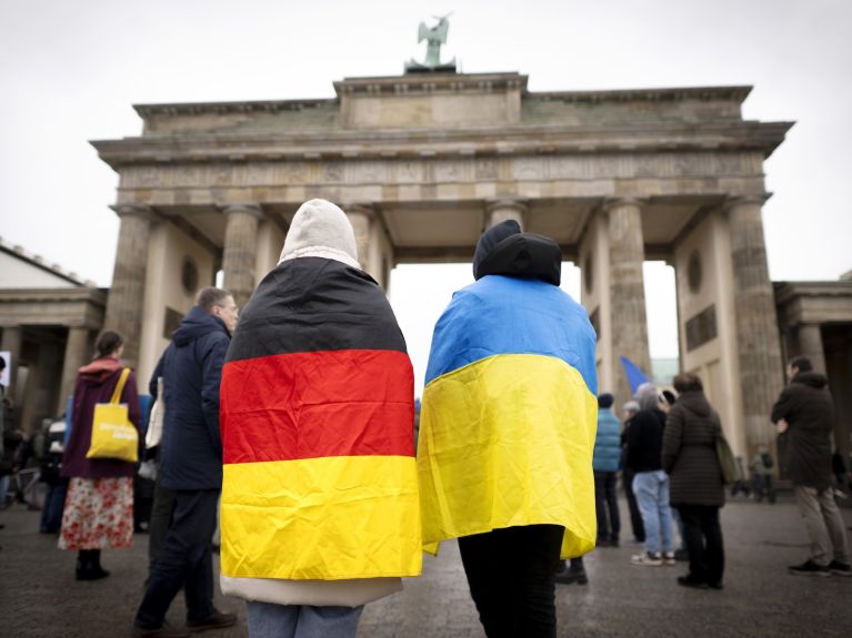 Eine Person mit einer Deutschland- und eine Person mit einer Ukraine-Flagge um die Schulter stehen vor dem Brandenburger Tor in Berlin