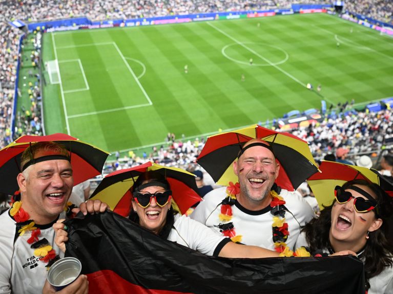 Vier Fußballfans in schwarz-rot-goldenen Hüten und mit Deutschland-Fanartikeln jubeln auf der Tribüne eines großen Stadions.