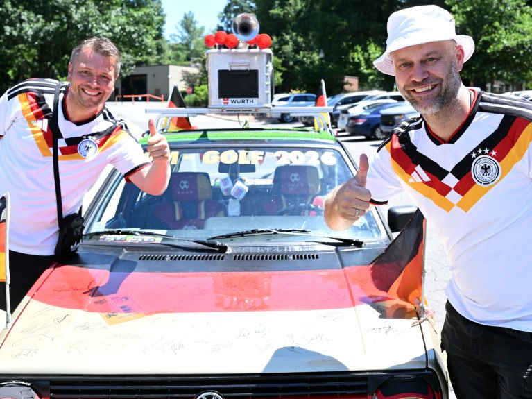 Zwei Männer in Deutschland-Trikots stehen lächelnd mit erhobenen Daumen neben einem schwarz-rot-goldenen VW Golf mit der Aufschrift „WM Golf 2026“.