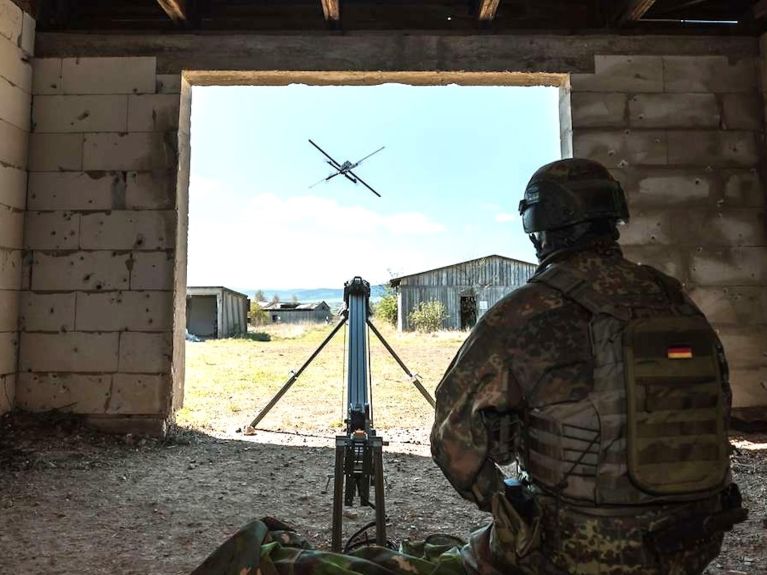 Soldat in Tarnuniform sitzt in einem Gebäude und steuert eine Drohne, die durch eine Maueröffnung ins Freie fliegt.