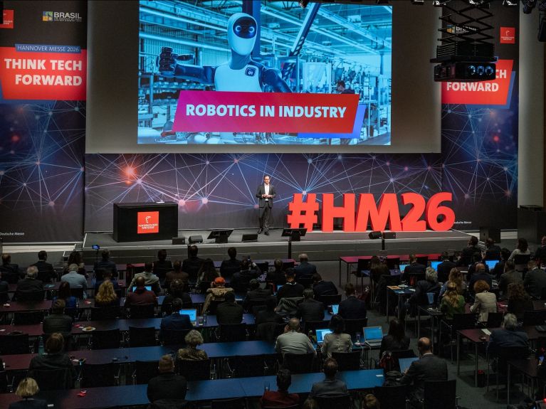 Ein Mann steht auf einer Bühne. Hinter ihm an einer Leinwand ist groß der Schriftzug „Robotics in Industry“ zu lesen.