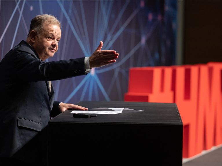 Ein älterer Mann steht auf einer Bühne an einem Rednerpult, neben ihm der Schriftzug „HM26“.