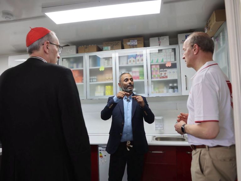 Ein Kardinal, ein Mann in Anzug und ein Mitarbeiter der Malteser in einem Behandlungszimmer