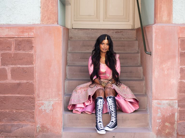 A young woman with Indian forehead jewellery and an opulent pink silk dress sits on the steps of a house entrance.