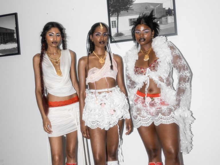 Three young women stand next to each other in relatively extravagant outfits consisting mainly of white lace and tulle.