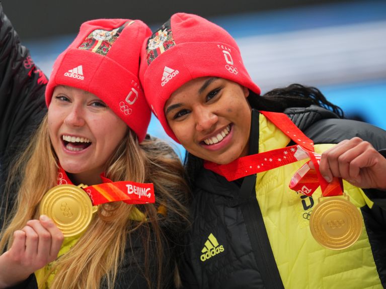 Портрет бобслеисток Лауры Нольте и Деборы Леви, склонивших головы друг к другу и держащих перед камерой свои золотые медали, завоеванные на зимних Олимпийских играх 2022 года в Пекине. 