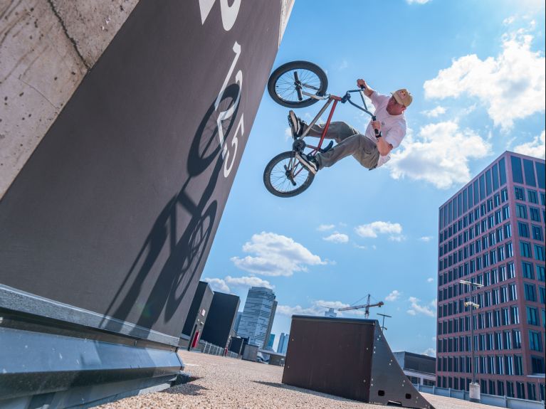 BMX-Fahrer springt mit dem Rad an einer Wandrampe hoch; im Hintergrund moderner Bürobau unter blauem Himmel.