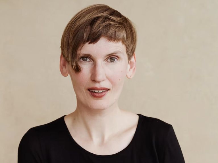 Portrait photo of a person with short hair wearing a black top against a light, blurred background who is looking directly into the camera. 