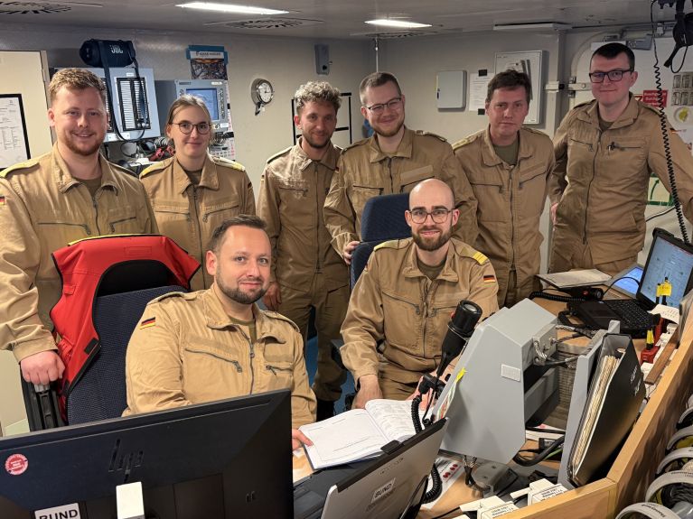 Two soldiers are sitting at a desk with a screen. Six further soldiers are standing behind them.  
