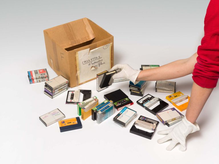 Arme einer Person, die weiße Handschuhe trägt und Audiokassetten sortiert, die vor einem Pappkarton liegen