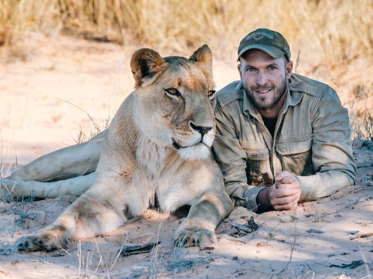 A man lies next to a lioness and smiles at the camera
