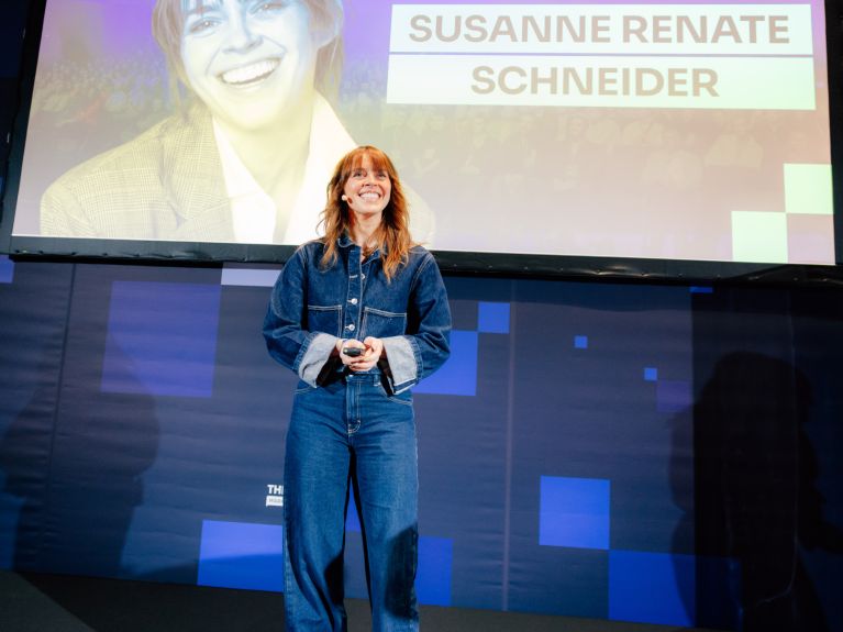 A smiling woman wearing a headset microphone stands on a stage holding a presentation remote, while her name, “SUSANNE RENATE SCHNEIDER”, is displayed on a large screen behind her.
