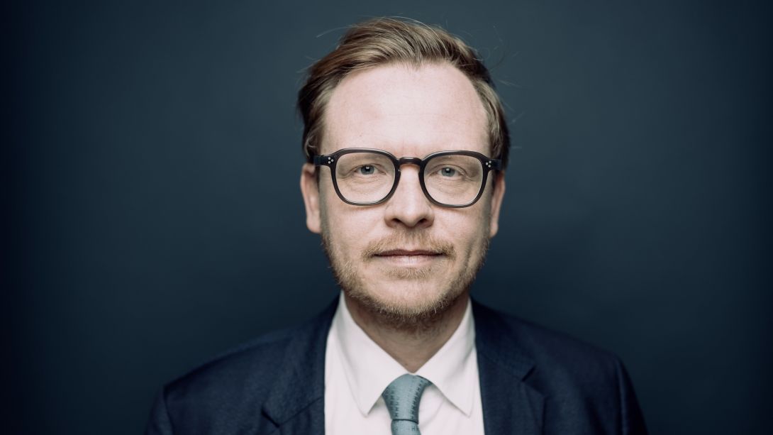 Porträt eines Mannes mit runder schwarzer Brille, kurzem Bart und Seitenscheitel, der in Anzug, weißem Hemd und hellgrüner Krawatte vor dunkelblauem Hintergrund in die Kamera blickt. 