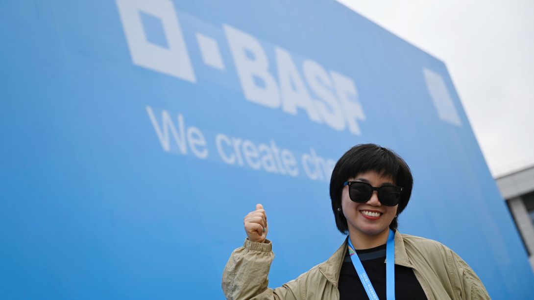 Chinesische Frau mit Sonnebrille zeigt auf eine blaue Wand mit BASF Logo hinter sich. 